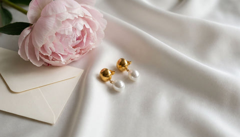Gold and white pearl drop earrings on folded white silk beside a pale pink peony and a plain cream envelope