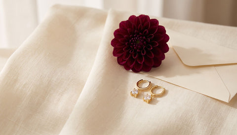 Gold huggie hoop earrings with a small clear stone on folded cream linen beside a burgundy dahlia and a plain cream envelope