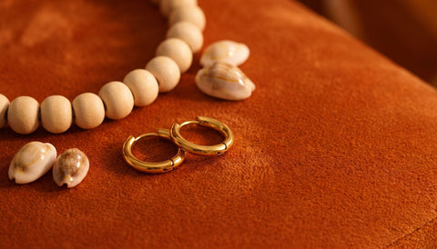 Small gold huggie hoop earrings on rust orange velvet beside a strand of wooden beads and small shells in warm light