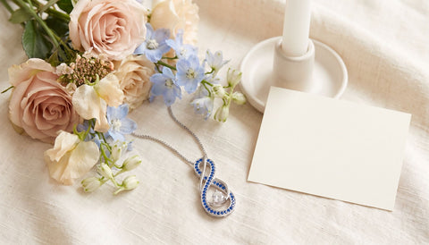 Blue stone infinity pendant necklace on cream linen beside garden flowers, an unlit white candle and a blank ivory card