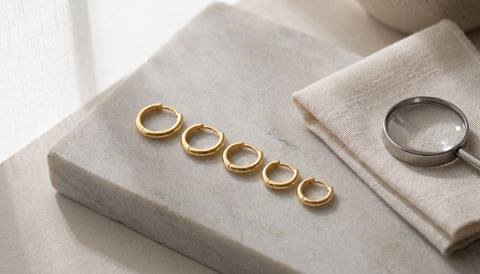 Five gold huggie earrings in graduated sizes on a pale stone slab beside a linen cloth and a magnifying loupe
