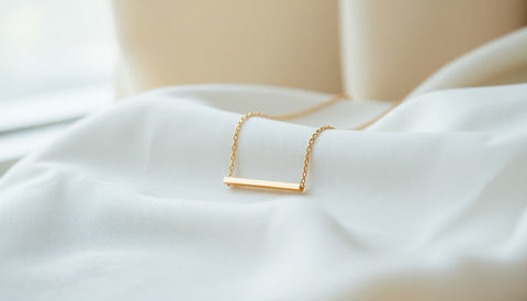 A delicate gold chain necklace with a slim horizontal bar pendant resting on soft neutral linen in natural light