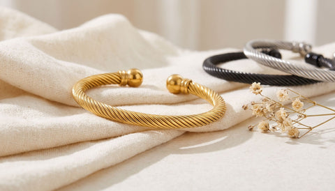 Gold twisted cable cuff bracelet on folded cream linen with a black and a silver cuff behind it and a sprig of dried flowers