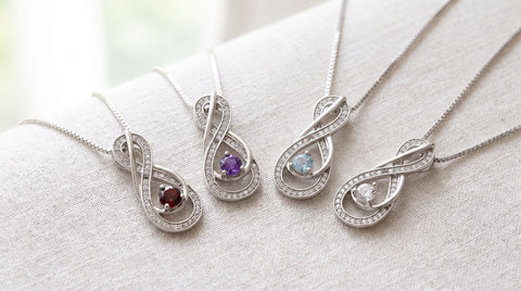 Four sterling silver birthstone infinity necklaces with red, purple, blue and clear stones arranged on a light surface