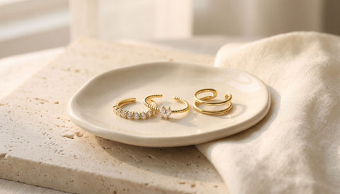 Three thin gold rings with small clear stones resting on a cream ceramic dish on pale stone beside a folded ivory cloth