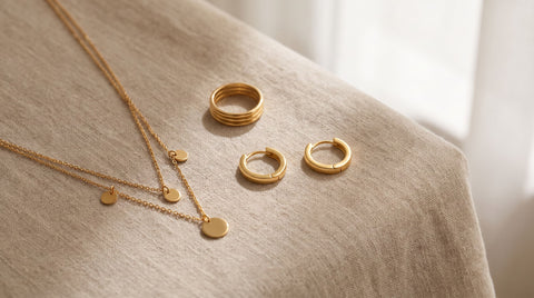 Flat-lay of everyday jewelry pieces including gold hoop earrings, a layered necklace and a stacking ring on linen fabric