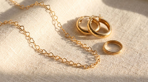 Assorted gold-plated jewelry pieces including a necklace, hoop earrings, and a ring arranged on a cream fabric surface