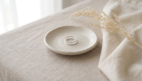 Thin silver double band ring resting on a small pale ceramic dish on natural linen beside dried grass and soft window light