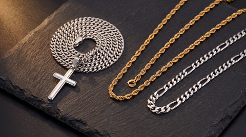Men's silver cuban link cross necklace coiled beside gold and silver chain necklaces on dark slate