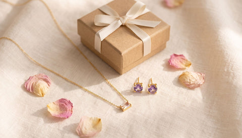 Gold birthstone pendant necklace and matching birthstone stud earrings styled as a Mother's Day gift flat-lay beside a wrapped kraft gift box