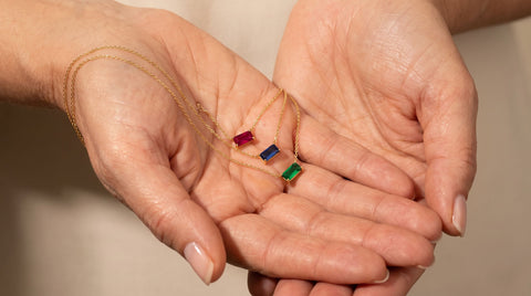 Three gold birthstone necklaces with ruby, sapphire, and emerald stones layered in an older woman's cupped hands