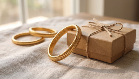 Gold stretch elastic bangle bracelet resting on neutral linen next to a small kraft gift box, with two more bracelets softly out of focus, styled as a bracelet gift for a daughter