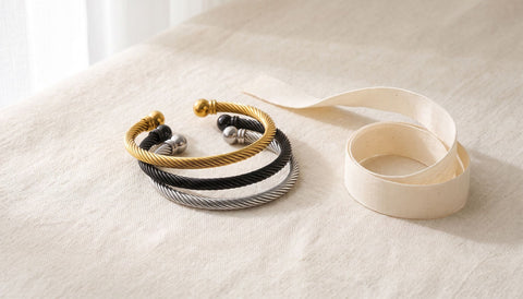 Open cuff bracelets in gold, black and silver tones beside a loop of plain cotton ribbon on cream linen