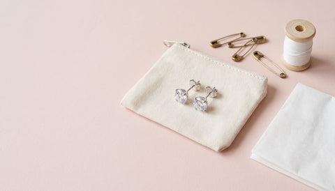 Pair of silver cubic zirconia stud earrings on a small cream zip pouch beside brass safety pins, a spool of white thread and a folded tissue on a blush surface