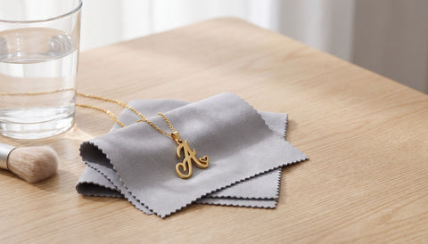 Gold script initial pendant necklace on a soft grey cloth on a light wood table beside a glass of water and a small soft brush