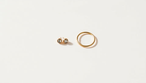 A pair of small round stud earrings and a tiny huggie hoop resting on a plain off white surface in soft daylight