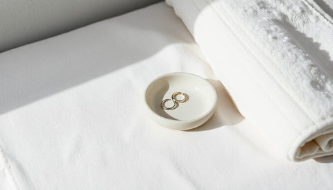 A small dish holding a pair of stud earrings and a thin ring on a folded linen cloth beside a soft towel