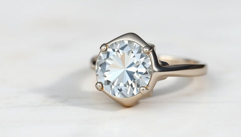 A solitaire ring with arched shoulders rising to a round clear stone, shown from the side on a soft stone surface