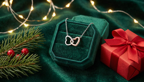 Two-tone silver and rose gold interlocking hearts necklace displayed in its gift box beside a wrapped red Christmas present, pine branches and warm string lights
