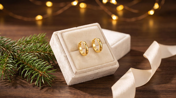 Gold huggie earrings in a cream velvet gift box beside evergreen and warm Christmas lights