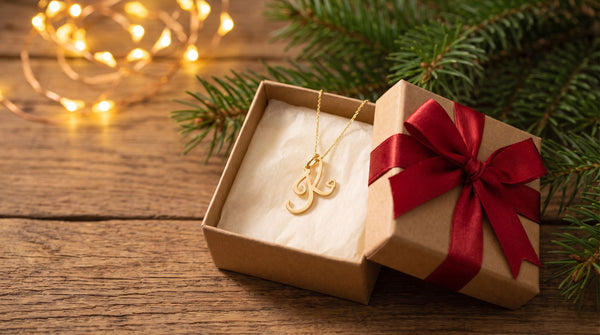 Gold script initial necklace in an open kraft gift box with a red ribbon, pine branches and warm lights