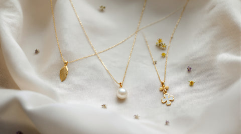 Delicate freshwater pearl necklace styled with a gold leaf charm and clover pendant on linen with dried wildflowers, cottagecore jewelry aesthetic flat lay