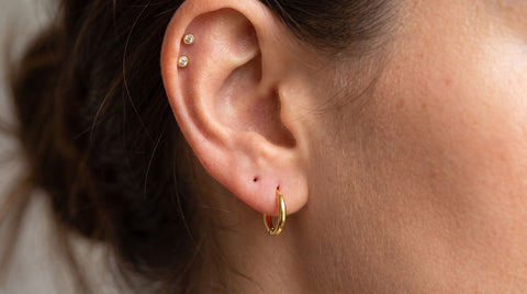 Close-up of a woman's ear showing correctly placed lobe and cartilage piercings with gold jewelry, illustrating an ear piercing chart