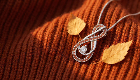 A silver infinity pendant necklace with orange-red stones resting on a rust orange knit beside two dried golden leaves