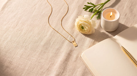 Gold initial tag necklace laid on linen beside a candle, a flower, and an open journal