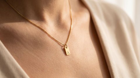 Simple gold initial tag pendant necklace worn at the collarbone, soft natural light