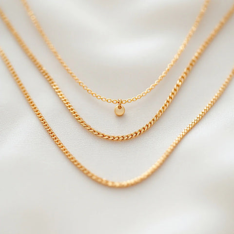 Three layered gold necklaces of different lengths and chain textures on a cream linen flat-lay