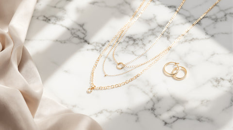 Layered gold-tone chain necklaces and small hoop earrings laid flat on marble for an Amyo Jewelry legitimacy review