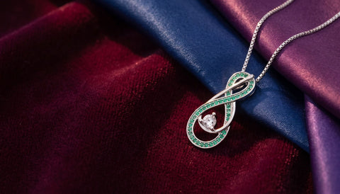 Silver infinity pendant necklace with green stones laid on deep burgundy velvet beside folded blue and purple silk