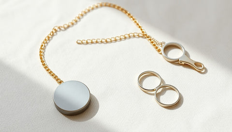 A small round magnet resting beside a gold chain, a silver ring and a plain metal clasp on a pale linen surface