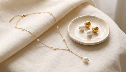 Fine gold necklace with small pearls and a pair of gold and pearl drop earrings in a cream ceramic dish on folded linen