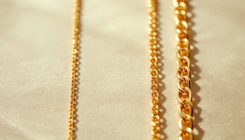 Three straight segments of plain gold chain of increasing thickness laid side by side on cream linen