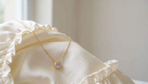 A thin gold necklace with a small round clear stone pendant resting on draped ivory satin with a softly ruffled edge