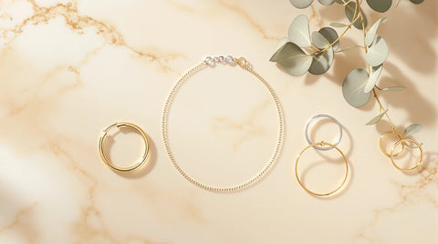 Nickel-free jewelry flat lay — sterling silver chain, 18K gold huggie earrings, and stacking rings on cream marble
