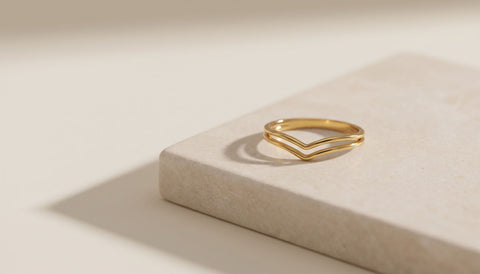 A thin gold chevron ring resting on a smooth pale beige stone slab with a soft side shadow