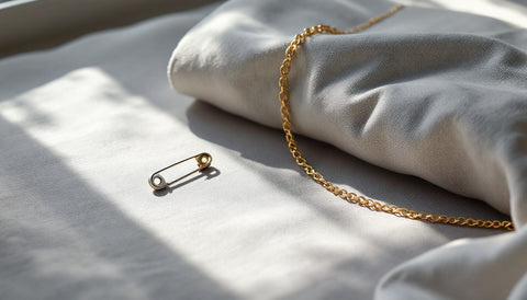 A single small safety pin on pale grey fabric beside a thin gold chain in soft window light