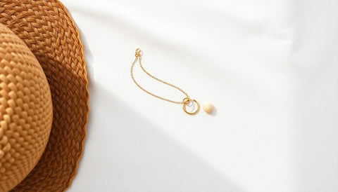 Gold anklet, small hoop earrings and a shell pendant laid on a sunlit linen cloth beside a straw hat