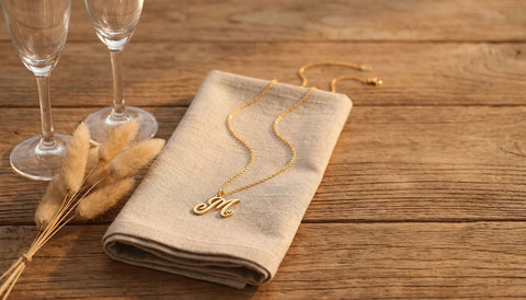 Gold script initial pendant necklace on a folded linen napkin on a wooden table beside two empty champagne flutes and dried grass stems