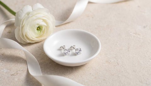 Pair of silver cubic zirconia stud earrings in a small white porcelain dish beside a white ranunculus flower and a loose satin ribbon on pale stone