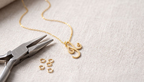 Fine gold chain with a script initial pendant on pale linen beside small smooth-jawed pliers and a few tiny loose gold rings