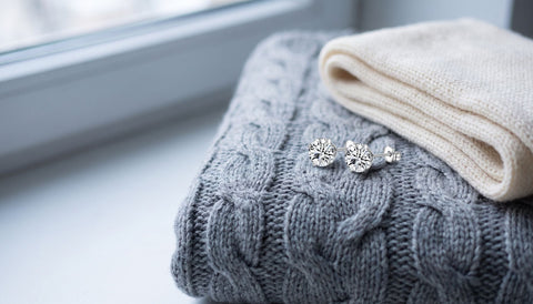 A pair of silver prong-set clear stone stud earrings on a grey cable-knit sweater beside a folded cream scarf