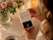Woman opening AJLuxe jewelry gift box with gold initial necklace beside flowers and candlelight for a thoughtful gift for mom